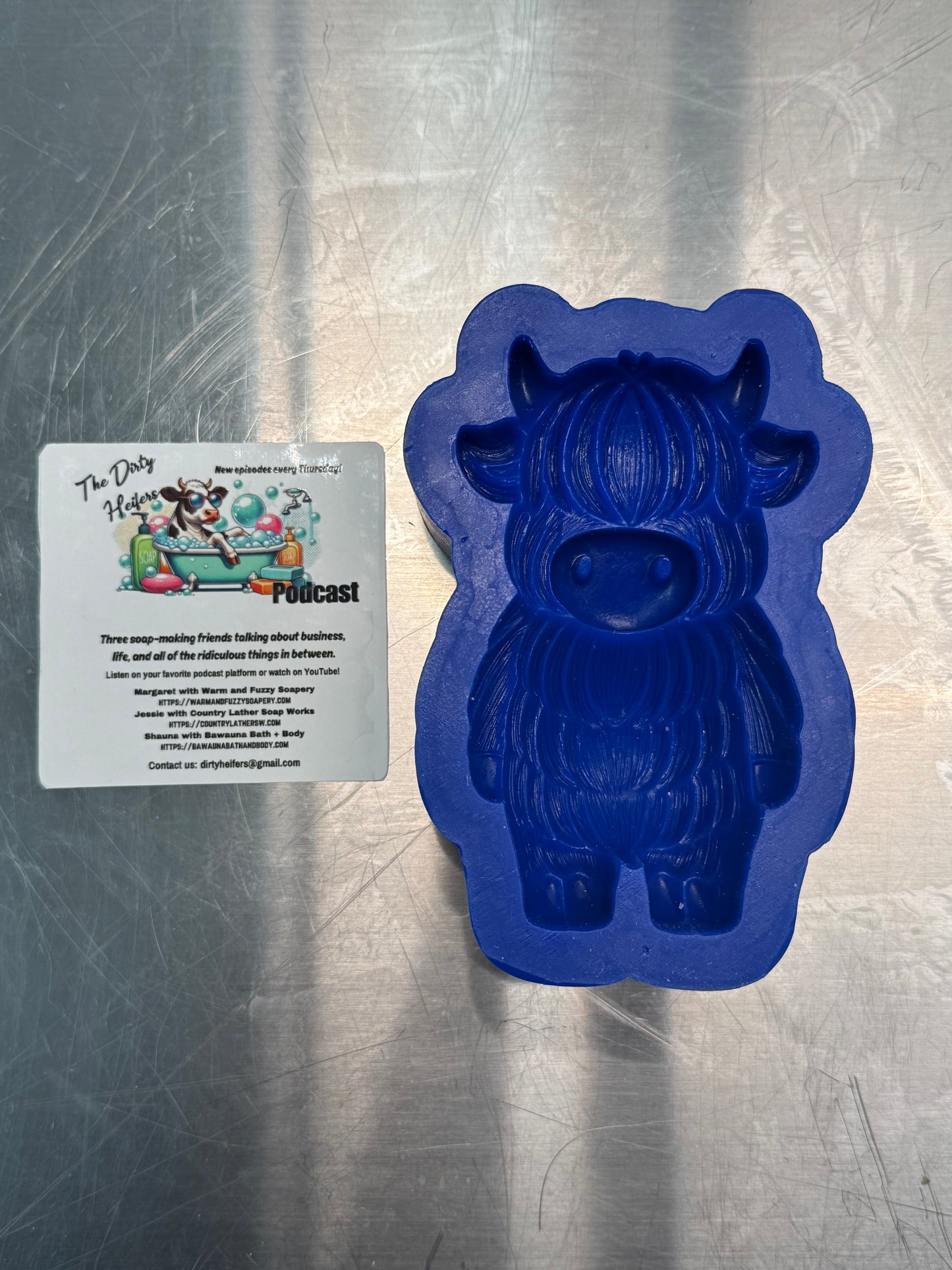 Blue silicone mold shaped like a cow on a metallic surface with a sticker for a dirty heifers podcast next to  mold
