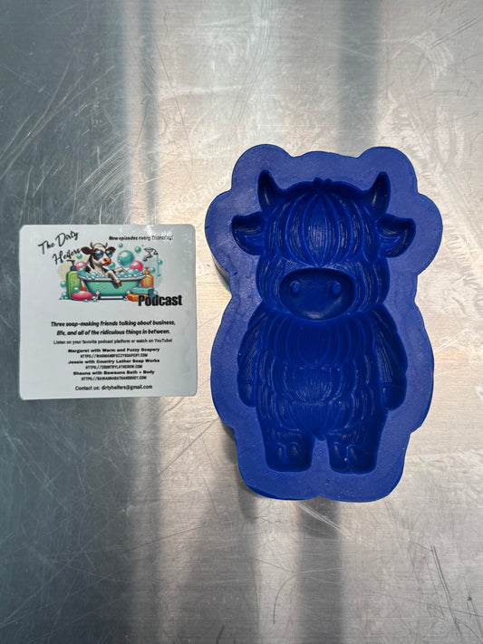 Blue silicone mold shaped like a cow on a metallic surface with a sticker for a dirty heifers podcast next to  mold